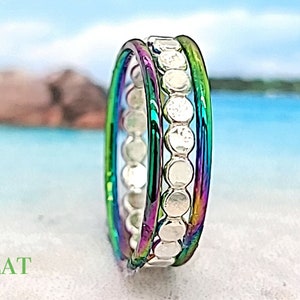 May include: A rainbow-colored ring with a silver beaded band. The ring is made of metal and has a flat, wide design.  FLAT BEADED & RAINBOW is written in green text on the white sand.