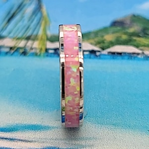 May include: A silver ring with a pink opal inlay. The ring is set against a tropical background.