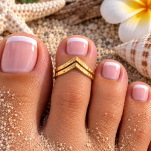 May include: A gold-toned metal toe ring featuring a double chevron or V-shaped design with a textured, hammered finish. The ring is worn on the second toe, which has a French manicure with a pale pink base and a crisp white tip.