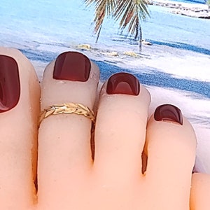 May include: Gold toe ring with a braided design. The ring is on a person's toe with dark red nail polish.