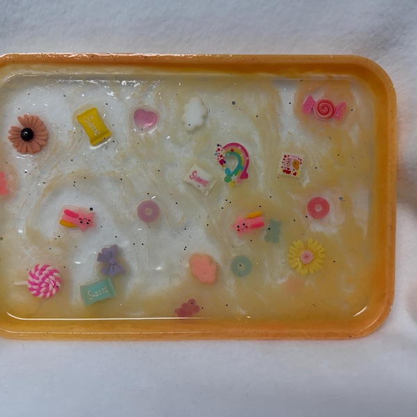 May include: A rectangular resin tray with a translucent white and yellow swirled base, featuring embedded miniature decorative charms including colorful candies, flowers, a rainbow, and small geometric shapes, all encased in a glossy finish.