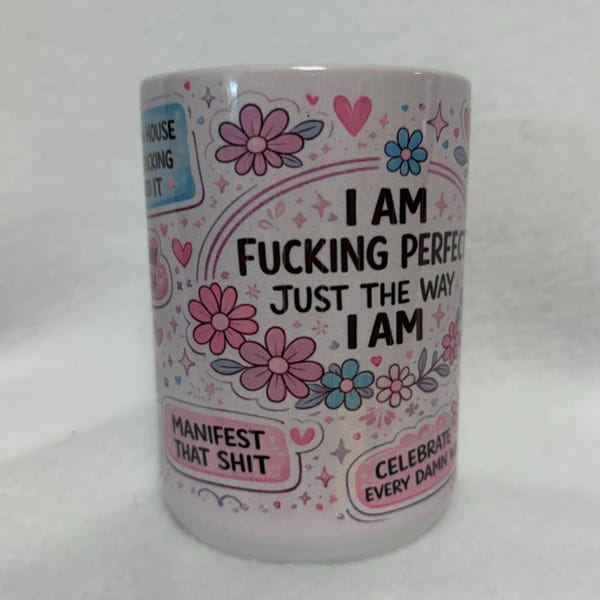 May include: A white ceramic mug featuring a pink and blue floral pattern with black text that reads I AM FUCKING PERFECT JUST THE WAY I AM, MANIFEST THAT SHIT, and CELEBRATE EVERY DAMN WIN, accented with small pink hearts and star shapes throughout.