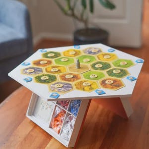 May include: A white wooden board game table with a hexagonal top. The game board features colorful hexagonal tiles and a central wooden piece. The table has a storage compartment underneath, holding game pieces.