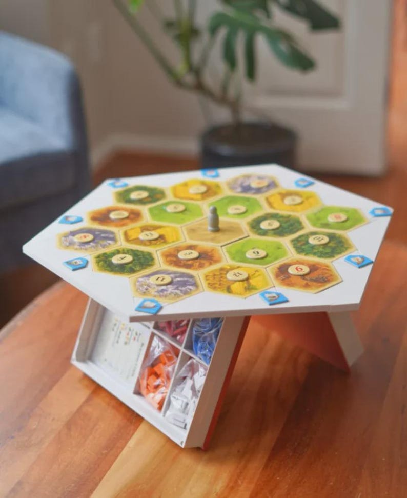 May include: A white wooden board game table with a hexagonal top. The game board features colorful hexagonal tiles and a central wooden piece. The table has a storage compartment underneath, holding game pieces.