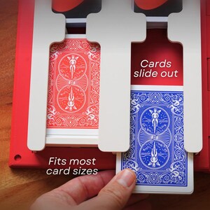 May include: A red and white card holder with two slots, each holding a deck of playing cards. One deck is red, the other blue. Text on the holder reads "Fits most card sizes" and "Cards slide out". A hand is holding the blue deck.