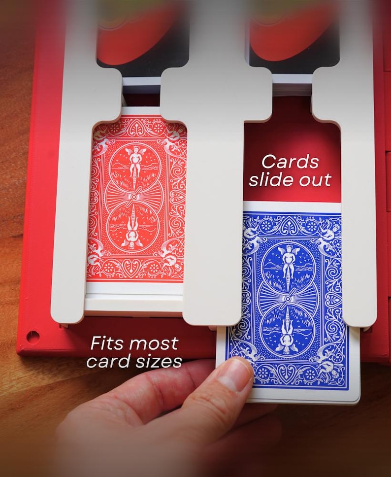 May include: A red and white card holder with two slots, each holding a deck of playing cards. One deck is red, the other blue. Text on the holder reads "Fits most card sizes" and "Cards slide out". A hand is holding the blue deck.