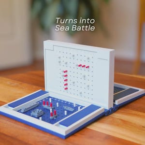 Op de afbeelding: Een wit en blauw Sea Battle-bord, open en klaar om te spelen. Het spelbord heeft een blauw speeloppervlak met miniatuur scheepsmodellen en rode pinnen. De tekst "Turns into Sea Battle" is zichtbaar.