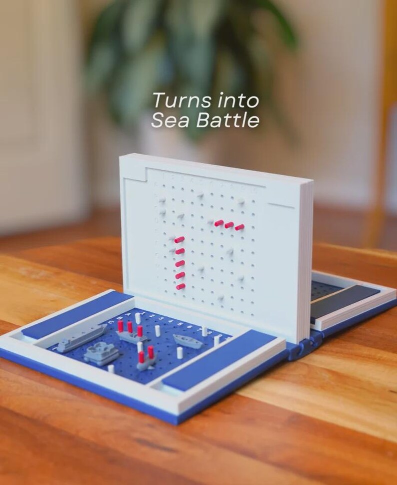 Op de afbeelding: Een wit en blauw Sea Battle-bord, open en klaar om te spelen. Het spelbord heeft een blauw speeloppervlak met miniatuur scheepsmodellen en rode pinnen. De tekst "Turns into Sea Battle" is zichtbaar.
