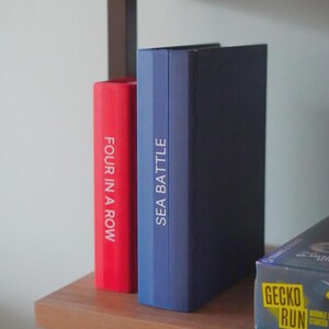 Op de afbeelding: Een houten plank toont twee bordspeldozen. E&eacute;n is rood met de tekst "FOUR IN A ROW" en de andere is blauw met "SEA BATTLE". Een derde doos met de tekst "GECKO RUN" is gedeeltelijk zichtbaar.