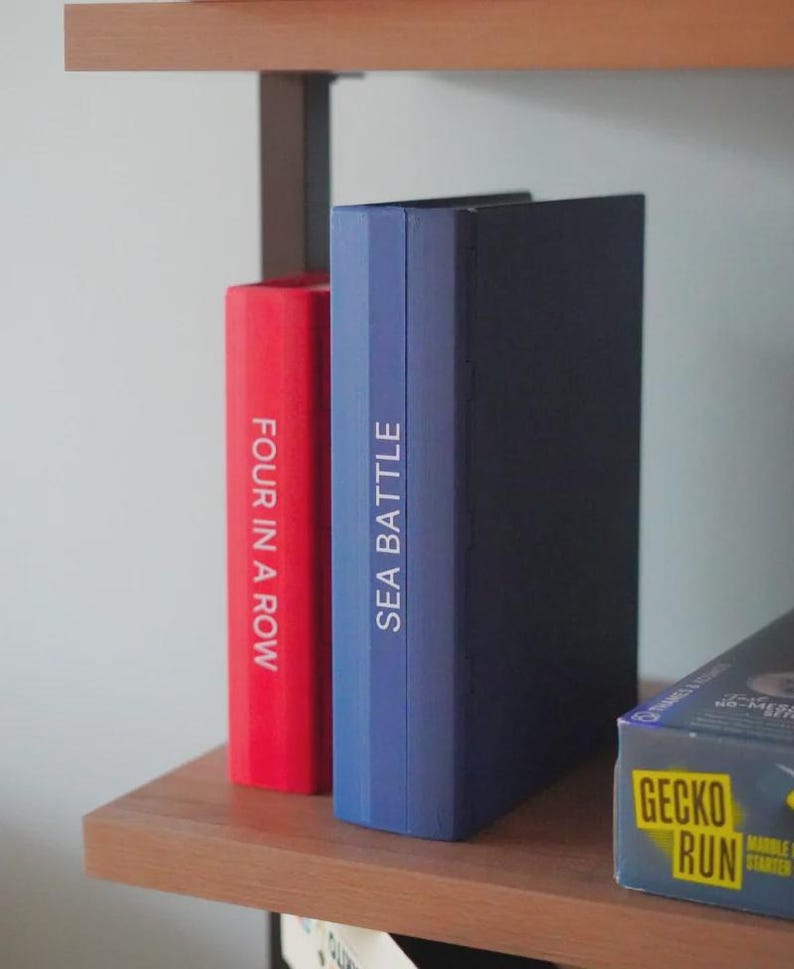 Op de afbeelding: Een houten plank toont twee bordspeldozen. E&eacute;n is rood met de tekst "FOUR IN A ROW" en de andere is blauw met "SEA BATTLE". Een derde doos met de tekst "GECKO RUN" is gedeeltelijk zichtbaar.