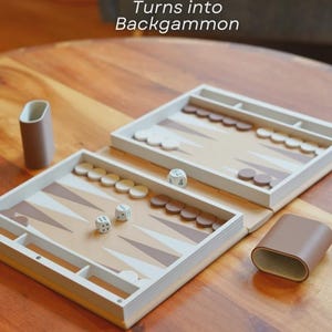 Puede incluir: Un juego de backgammon con una superficie de juego marr&oacute;n claro y blanca, dados y fichas. El texto "Turns into Backgammon" es visible. El juego est&aacute; abierto sobre una mesa de madera, con un estuche marr&oacute;n.