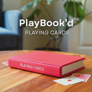 May include: A red playing card case with the words "PLAYING CARDS" in white lettering. The case is on a wooden table, with two playing cards visible. The text "PlayBook'd" and "PLAYING CARDS" are also visible.