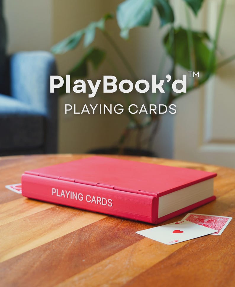 May include: A red playing card case with the words "PLAYING CARDS" in white lettering. The case is on a wooden table, with two playing cards visible. The text "PlayBook'd" and "PLAYING CARDS" are also visible.