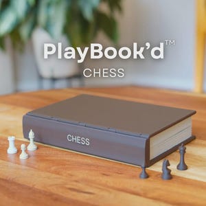 May include: A closed, brown chess set in the shape of a book, with the word "CHESS" printed on the side. White and black chess pieces are arranged on a wooden surface. The text "PlayBook'd" is at the top.