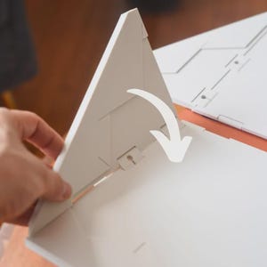 May include: A white, geometric, folded paper structure with a triangular shape. The structure is being opened, revealing its interior. A white arrow indicates the direction of the opening. The background is a warm, neutral color.