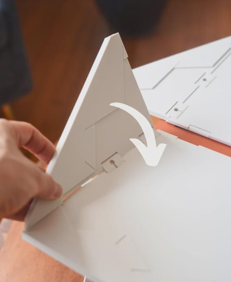 May include: A white, geometric, folded paper structure with a triangular shape. The structure is being opened, revealing its interior. A white arrow indicates the direction of the opening. The background is a warm, neutral color.