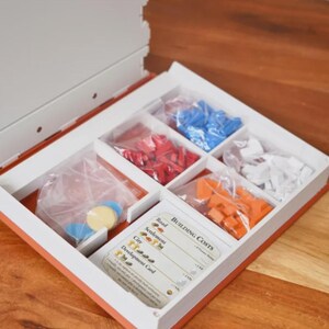 May include: Open board game box with compartments containing red, blue, orange, and white game pieces. The box also includes cards with building costs and game instructions. The box is white with an orange trim.