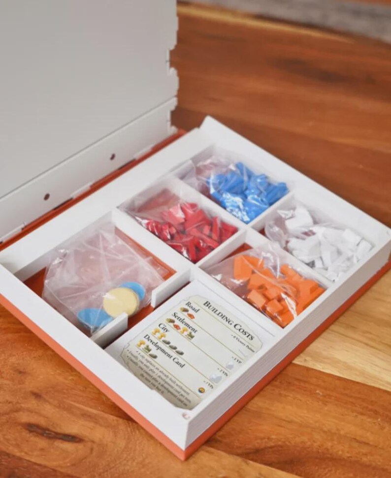 May include: Open board game box with compartments containing red, blue, orange, and white game pieces. The box also includes cards with building costs and game instructions. The box is white with an orange trim.