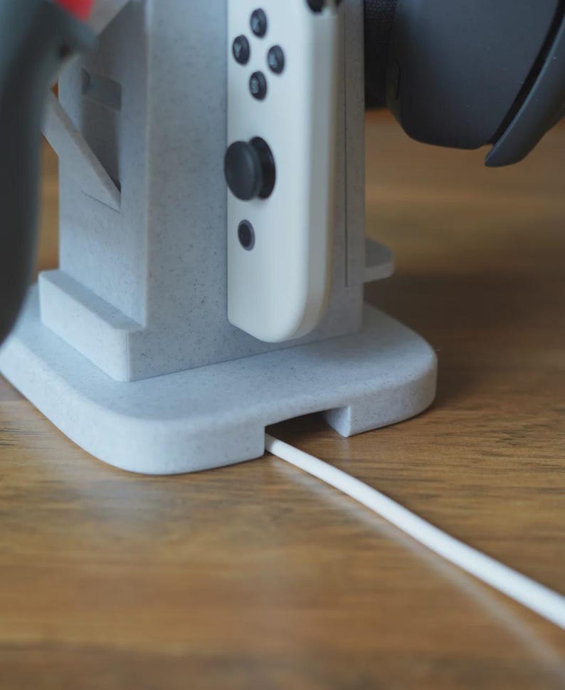 Może przedstawiać: Jasnoszary, drukowany w 3D stojak na konsolę do gier. Biały kontroler jest zadokowany na stojaku, z białym kablem wychodzącym z podstawy. Czarny zestaw słuchawkowy spoczywa na stojaku. Stojak stoi na drewnianej powierzchni.