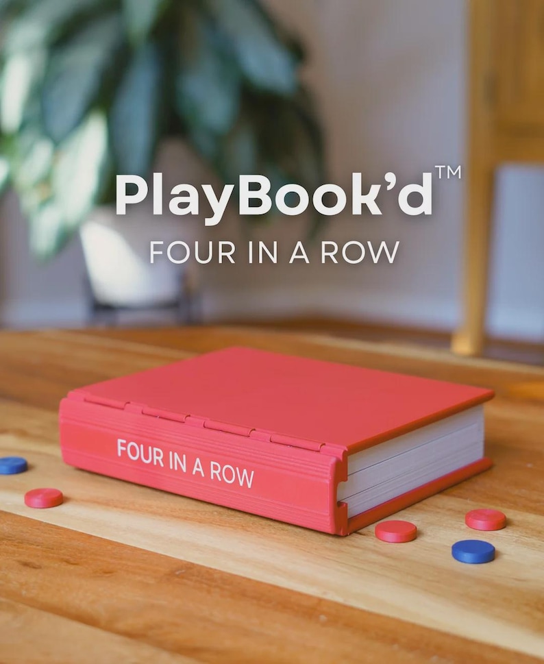 Może przedstawiać: Czerwona gra PlayBook'd Four in a Row na drewnianym stole. Gra jest w etui przypominającym książkę z białym tekstem. Rozrzucone są czerwone i niebieskie elementy gry.