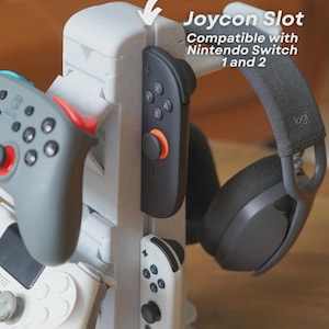 Może przedstawiać: Biało-szary stojak na konsolę do gier z wieloma gniazdami na kontrolery i zestaw słuchawkowy. Stojak zawiera tekst "Joycon Slot Compatible with Nintendo Switch 1 and 2". Widoczne są kontrolery w kolorze szarym, czarnym i białym.