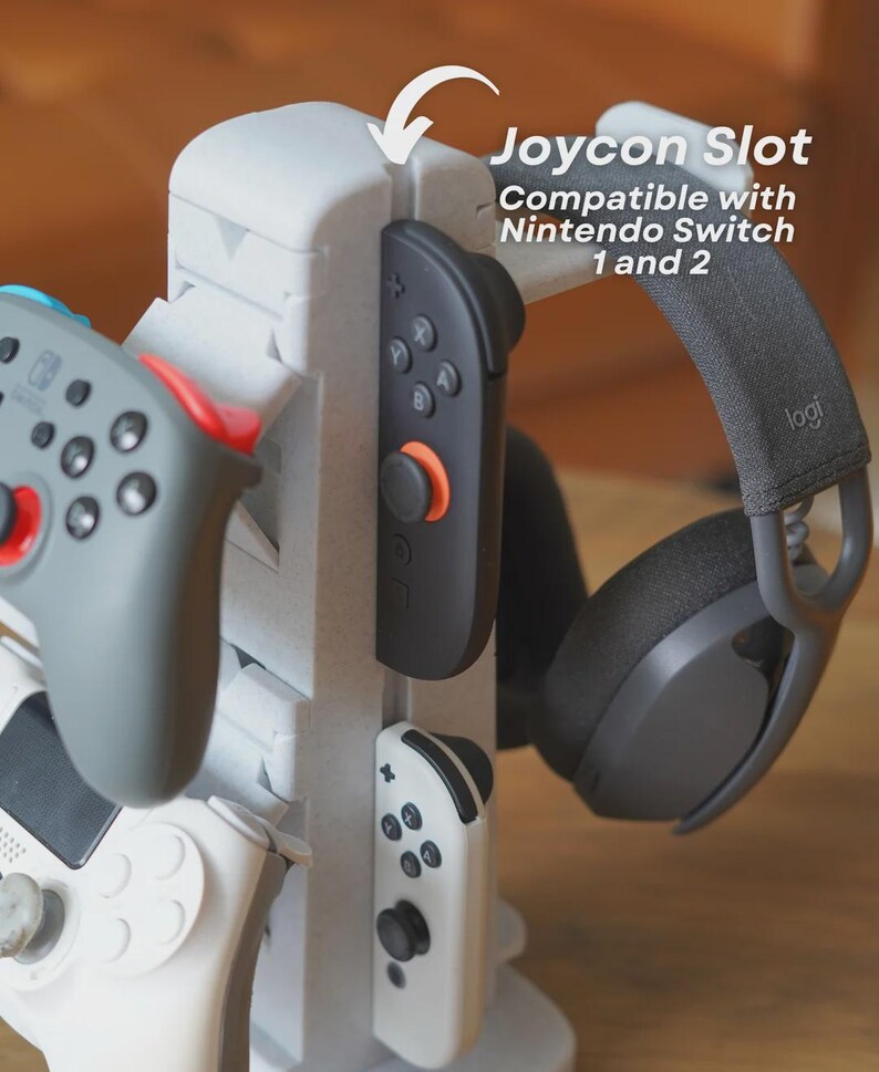 Może przedstawiać: Biało-szary stojak na konsolę do gier z wieloma gniazdami na kontrolery i zestaw słuchawkowy. Stojak zawiera tekst "Joycon Slot Compatible with Nintendo Switch 1 and 2". Widoczne są kontrolery w kolorze szarym, czarnym i białym.