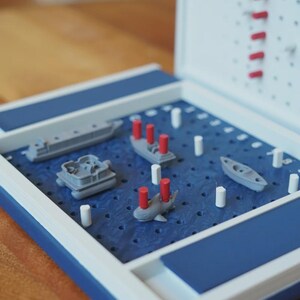 Op de afbeelding: Een Battleship-spelbord met een blauw speeloppervlak en een wit en blauw frame. Het spel bevat grijze miniatuurschepen en rode en witte pinnen. Het spel is open, klaar om te spelen.