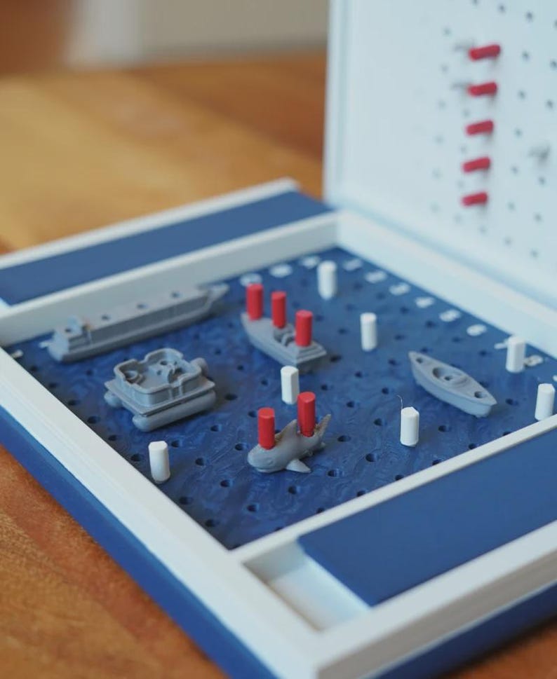 Op de afbeelding: Een Battleship-spelbord met een blauw speeloppervlak en een wit en blauw frame. Het spel bevat grijze miniatuurschepen en rode en witte pinnen. Het spel is open, klaar om te spelen.