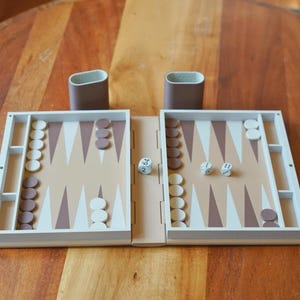 Puede incluir: Un juego de backgammon de madera con una superficie de juego beige claro, puntas triangulares marrones y blancas, y fichas blancas y marrones. Se ven dos dados en el tablero. Tambi&eacute;n hay dos portavasos marrones.
