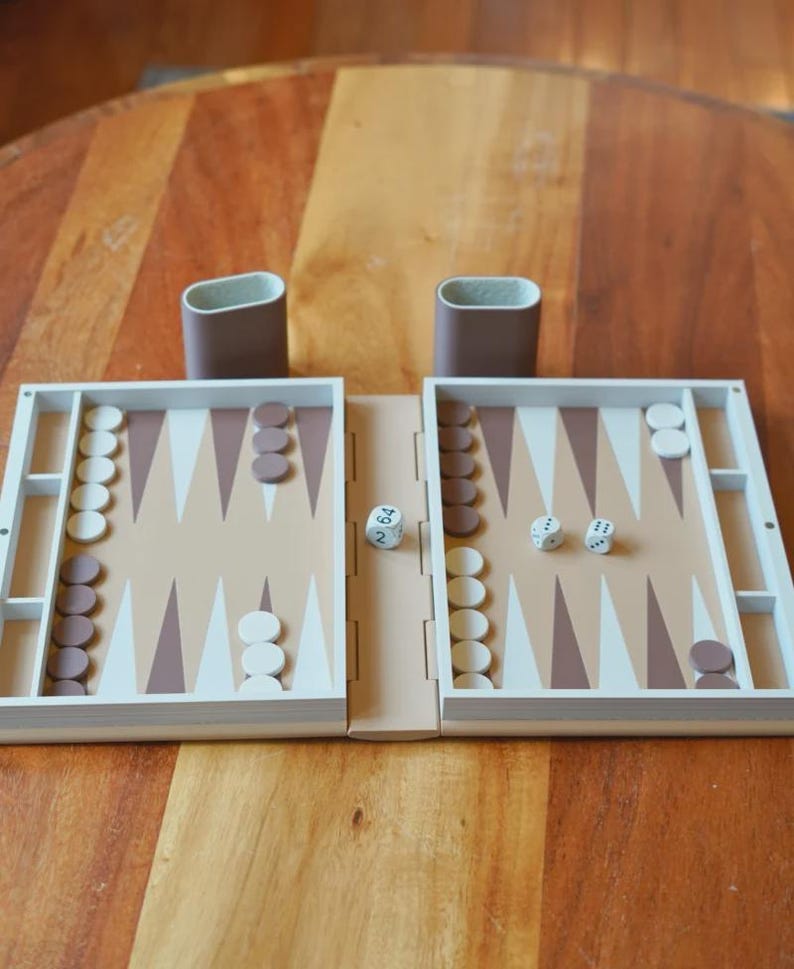 Puede incluir: Un juego de backgammon de madera con una superficie de juego beige claro, puntas triangulares marrones y blancas, y fichas blancas y marrones. Se ven dos dados en el tablero. Tambi&eacute;n hay dos portavasos marrones.