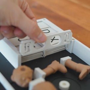 May include: A white plastic game with a black base. A hand is holding a white piece with a black "X" symbol. The game includes small, tan-coloured body parts and white rectangular pieces with letters.