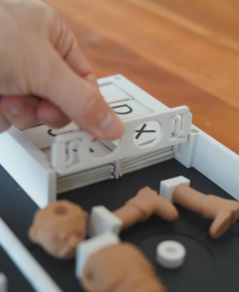 May include: A white plastic game with a black base. A hand is holding a white piece with a black "X" symbol. The game includes small, tan-coloured body parts and white rectangular pieces with letters.