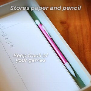May include: A white tray with a pink pen and a notepad. The notepad has handwritten numbers and the text "Keep track of your games" and "Stores paper and pencil". The pen is pink with a white tip.