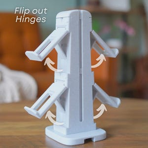 Może przedstawiać: Jasnoszare plastikowe urządzenie z odchylanymi zawiasami, przeznaczone do stania w pozycji pionowej na płaskiej powierzchni. Tekst "Flip out Hinges" jest widoczny w lewym g&oacute;rnym rogu. Zawiasy są pokazane w pozycji otwartej z zakrzywionymi strzałkami.