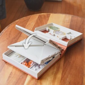 May include: Open board game box with white and orange accents. The box is open, revealing game pieces and cards. The lid is partially open, with an arrow indicating the opening direction. The box is on a wooden table.