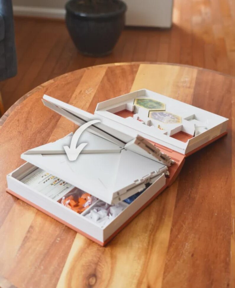 May include: Open board game box with white and orange accents. The box is open, revealing game pieces and cards. The lid is partially open, with an arrow indicating the opening direction. The box is on a wooden table.