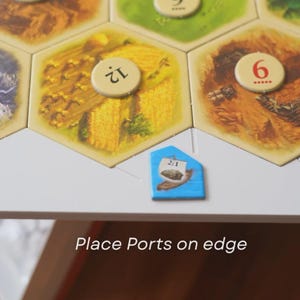 May include: A board game with hexagonal tiles depicting fields and resources. The game board includes a blue port tile with the text "2:1" and the instruction "Place Ports on edge". The tiles have numbers and illustrations of wheat, forest, and other resources.