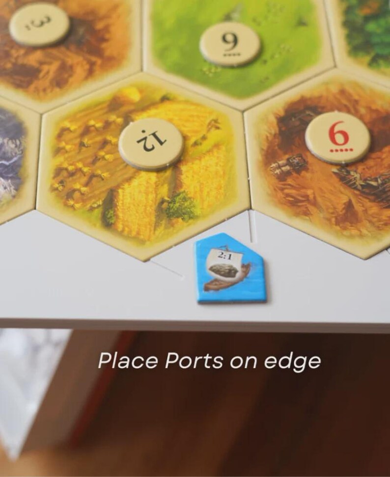 May include: A board game with hexagonal tiles depicting fields and resources. The game board includes a blue port tile with the text "2:1" and the instruction "Place Ports on edge". The tiles have numbers and illustrations of wheat, forest, and other resources.