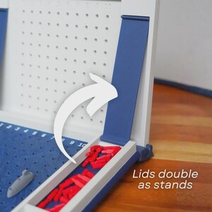 Op de afbeelding: Een wit en blauw Battleship-bordspel met rode spelstukken. Het spelbord heeft een blauw speeloppervlak met een grijs schip en een wit raster. De tekst "Lids double as stands" is zichtbaar.