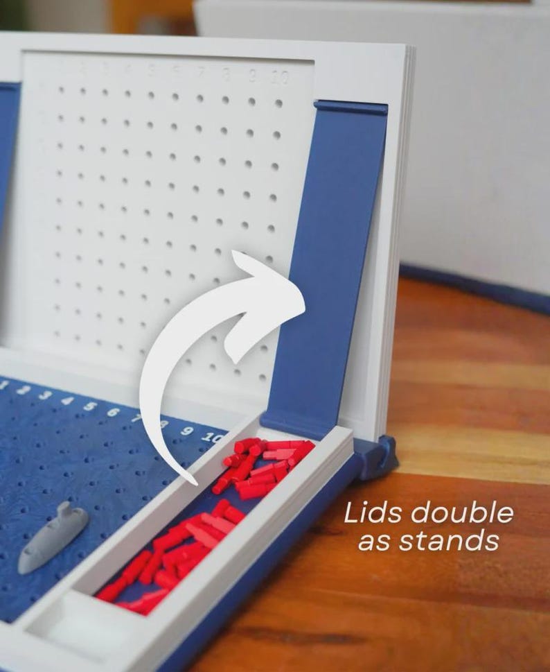 Op de afbeelding: Een wit en blauw Battleship-bordspel met rode spelstukken. Het spelbord heeft een blauw speeloppervlak met een grijs schip en een wit raster. De tekst "Lids double as stands" is zichtbaar.
