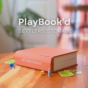May include: An orange Settlers' Storage box, designed to look like a book, with the text "PlayBook'd" and "SETTLERS' STORAGE". Dice, game pieces, and cards are on the wooden table. The box is rectangular with rounded edges.