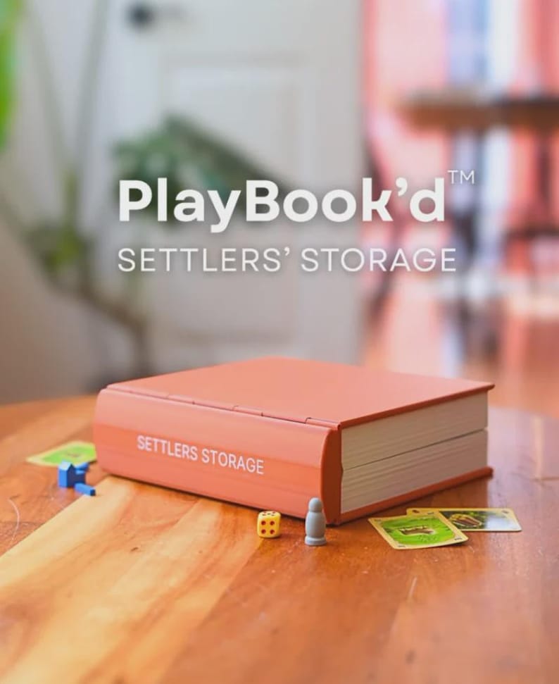 May include: An orange Settlers' Storage box, designed to look like a book, with the text "PlayBook'd" and "SETTLERS' STORAGE". Dice, game pieces, and cards are on the wooden table. The box is rectangular with rounded edges.