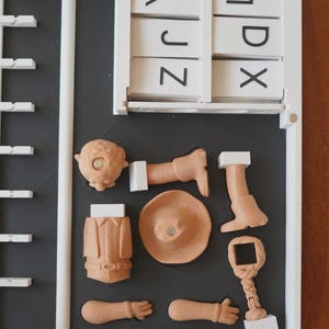 May include: A white framed puzzle with a black interior. The puzzle contains a set of white letter tiles and several tan-coloured puzzle pieces, including a head, arms, legs, a hat, and a jacket. The puzzle pieces are arranged in a specific order.
