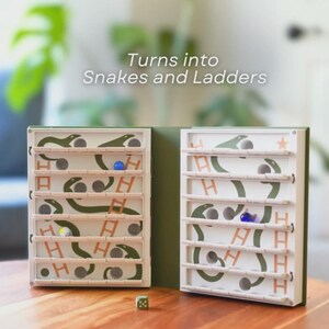 May include: A Snakes and Ladders game with a green and white design. The game board is open, revealing the playing surface with snakes, ladders, and spaces. The text "Turns into Snakes and Ladders" is visible. A die and game pieces are on the wooden table.