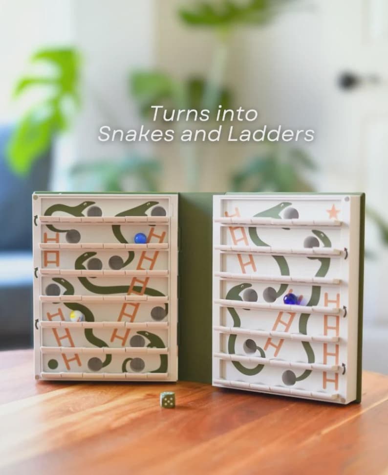 May include: A Snakes and Ladders game with a green and white design. The game board is open, revealing the playing surface with snakes, ladders, and spaces. The text "Turns into Snakes and Ladders" is visible. A die and game pieces are on the wooden table.