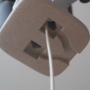 Może przedstawiać: Zbliżenie jasnobeżowego, teksturowanego, kwadratowego obiektu z białym przewodem wychodzącym z kwadratowego otworu. Obiekt ma rzeźbiony wz&oacute;r i jest umieszczony na miękkim, neutralnym tle.