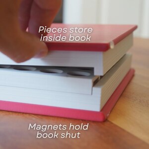 Może przedstawiać: Czerwono-biała książka z tekstem "Pieces store inside book" i "Magnets hold book shut". Książka jest otwarta, odsłaniając schowek. Książka leży na drewnianej powierzchni.