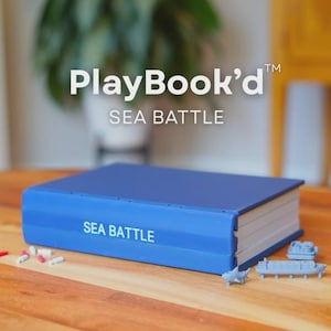 Op de afbeelding: Een blauwe PlayBook'd Sea Battle-spelset in de vorm van een boek. De voorkant toont de tekst "PlayBook'd" en "SEA BATTLE". De set bevat kleine speelgoed schepen en spelstukken. Het spel staat op een houten tafel.