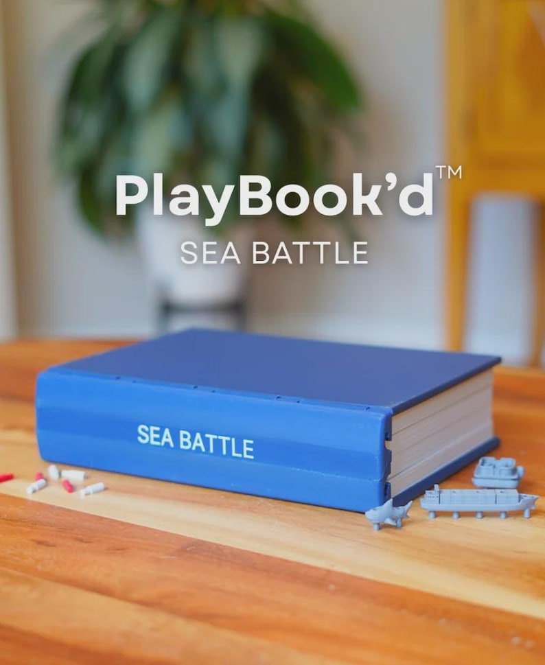 Op de afbeelding: Een blauwe PlayBook'd Sea Battle-spelset in de vorm van een boek. De voorkant toont de tekst "PlayBook'd" en "SEA BATTLE". De set bevat kleine speelgoed schepen en spelstukken. Het spel staat op een houten tafel.