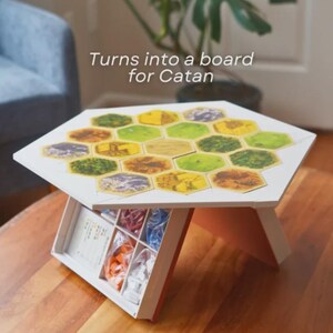 May include: A white hexagonal board game table with a colorful Catan game board design. The table has a built-in storage compartment with game pieces. The text "Turns into a board for Catan" is visible on the image.